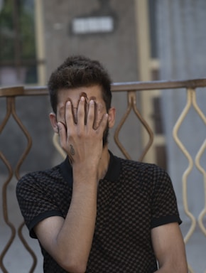 A person is seated with their hand covering their face, obscuring their identity. They have a tattoo on their wrist, and the background includes a patterned rail and a slightly blurred wall.