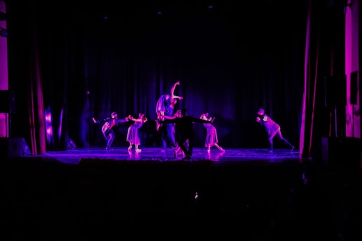 A vibrant dance rehearsal with a coach guiding a diverse group of performers under dramatic stage lighting.