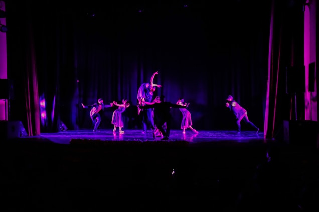A moody, cinematic shot of four dancers mid-move, bathed in neon purple and pink light against a deep black background.