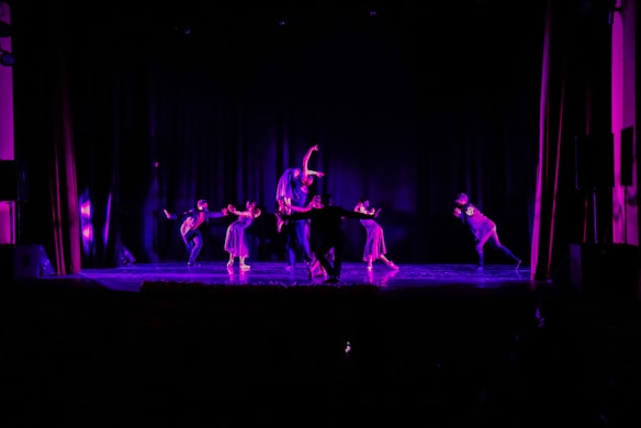 A group of dancers is performing on a dimly lit stage, bathed in purple and blue lighting. The setting is dramatic, with dancers in varying poses that convey motion and expression. The background is dark, highlighting the performers.