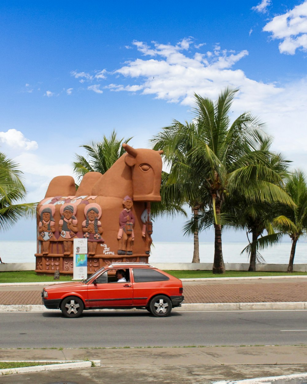 a red car driving past a large sculpture of elephants