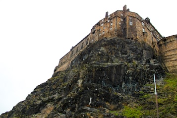 A historic castle perched atop a rocky hill, with its stone structure blending into the dark rock formation. The architecture features turrets and several small windows, indicative of a medieval fortress. The sky is overcast and the rocks are covered in patches of moss, adding to the dramatic appearance.