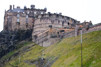 A historic stone castle is perched on a rocky hill, with massive walls and turrets. The slope leading up to the walls is covered in green grass. The castle features various architectural styles, showcasing a blend of medieval and Renaissance elements.