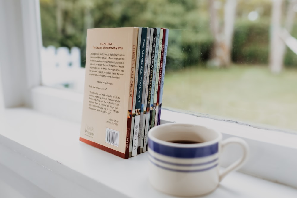 a stack of books sitting on top of a window sill next to a cup