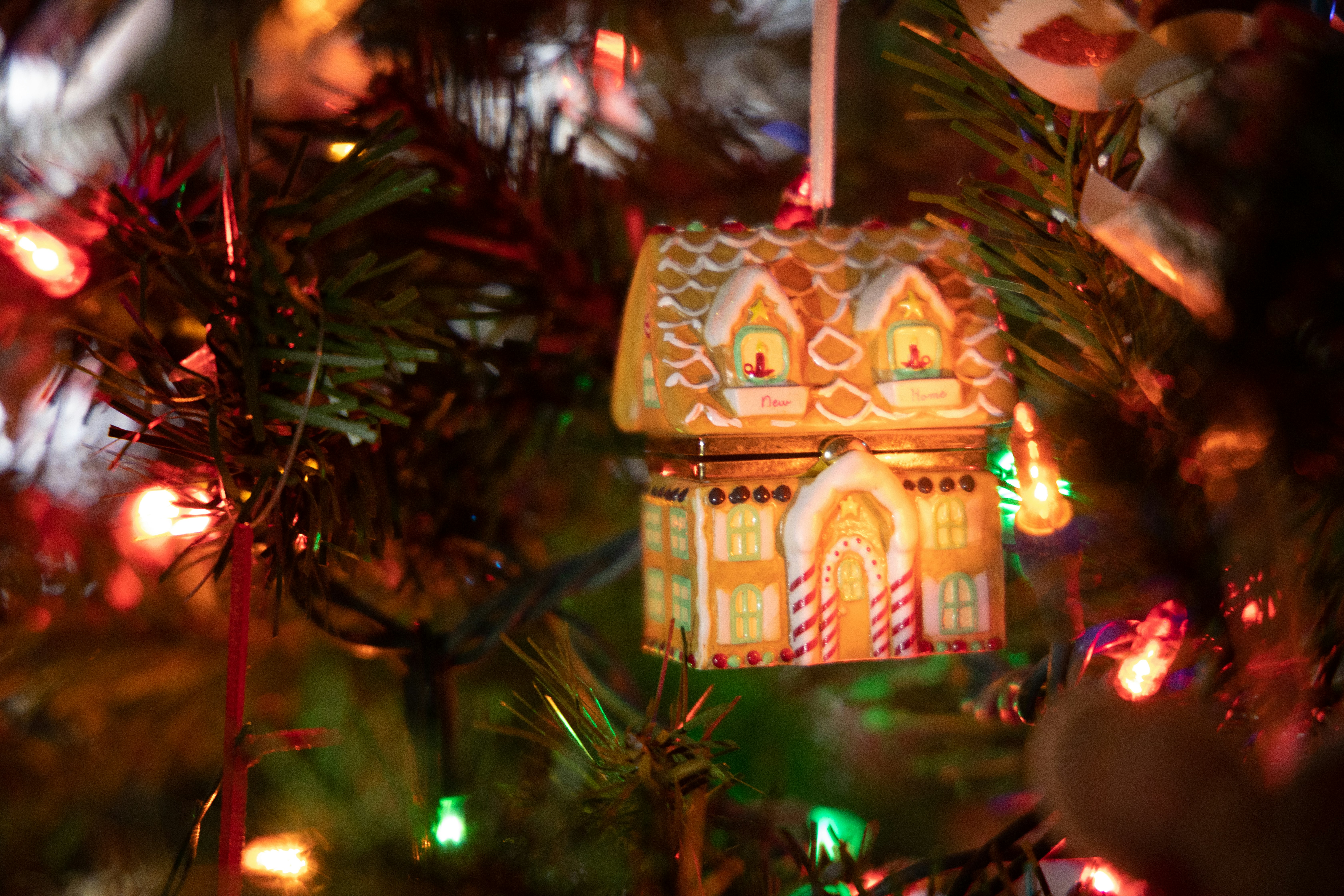 A small, house-shaped Christmas ornament hangs on a tree branch surrounded by multicolored holiday lights.