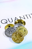 The image shows several cryptocurrency coins placed on a white surface with the word 'QUANT' partially visible in the background. There are both gold and silver colored coins featuring various designs and symbols representing different cryptocurrencies.