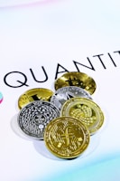 The image shows several cryptocurrency coins placed on a white surface with the word 'QUANT' partially visible in the background. There are both gold and silver colored coins featuring various designs and symbols representing different cryptocurrencies.
