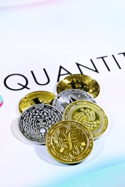 The image shows several cryptocurrency coins placed on a white surface with the word 'QUANT' partially visible in the background. There are both gold and silver colored coins featuring various designs and symbols representing different cryptocurrencies.