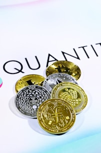 The image shows several cryptocurrency coins placed on a white surface with the word 'QUANT' partially visible in the background. There are both gold and silver colored coins featuring various designs and symbols representing different cryptocurrencies.