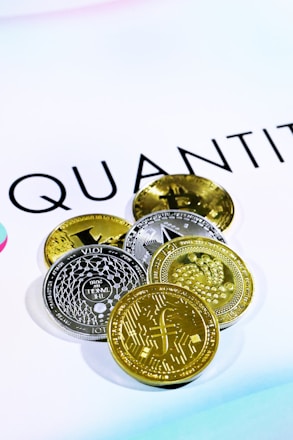 The image shows several cryptocurrency coins placed on a white surface with the word 'QUANT' partially visible in the background. There are both gold and silver colored coins featuring various designs and symbols representing different cryptocurrencies.