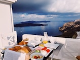 A variety of breakfast foods is arranged on a table with a picturesque view of a calm sea and distant islands under a cloudy sky. The meal includes bread rolls in a basket, sliced fruits, a glass of orange juice, and bowls of condiments. There is a white chair on the left side of the table, and the setting suggests a peaceful outdoor dining experience on a balcony or terrace.
