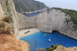 A secluded beach surrounded by steep cliffs with a rusting shipwreck on the sandy shore. The vibrant blue of the water contrasts with the tan and brown hues of the cliffs and the green vegetation atop them. Boats are anchored in the cove, and visitors are scattered along the beach.