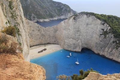A secluded beach surrounded by steep cliffs with a rusting shipwreck on the sandy shore. The vibrant blue of the water contrasts with the tan and brown hues of the cliffs and the green vegetation atop them. Boats are anchored in the cove, and visitors are scattered along the beach.
