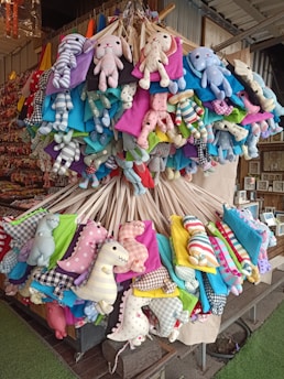 A collection of colorful plush toys, including animals and pillow-like shapes, is suspended in a cluster from a display. The toys feature various patterns, such as stripes and polka dots, and are made from a range of bright and pastel fabrics. The setting appears to be a market or shop with additional items visible in the background.