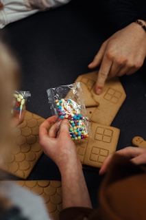 Hands are seen preparing gingerbread house pieces and holding a plastic bag filled with colorful candies. A person points at one of the gingerbread panels placed on a dark surface. The gingerbread pieces have embossed designs resembling windows and roof tiles.