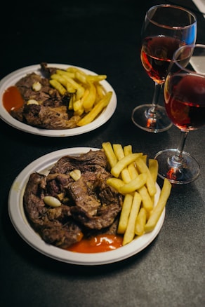 Two white plates hold servings of grilled steak and crispy golden fries, accompanied by a dollop of ketchup. Two glasses of red wine sit beside the plates, set against a dark background creating an intimate dining atmosphere.