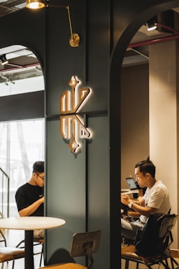 In a modern cafe setting, two people are seated across from each other, each focused on their devices. A stylish emblem is mounted on a dark-colored partition wall, with industrial-style lighting enhancing the ambiance.