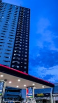 A tall residential building stands adjacent to a gas station, against a backdrop of a deep blue evening sky. The gas station has several fuel pumps visible under a well-lit overhang. Brand signage is prominently displayed on the pumps and the station roof, with a nearby shop with 'Treats' written on its signage.