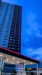 A tall residential building stands adjacent to a gas station, against a backdrop of a deep blue evening sky. The gas station has several fuel pumps visible under a well-lit overhang. Brand signage is prominently displayed on the pumps and the station roof, with a nearby shop with 'Treats' written on its signage.