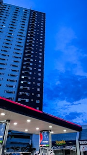 A tall residential building stands adjacent to a gas station, against a backdrop of a deep blue evening sky. The gas station has several fuel pumps visible under a well-lit overhang. Brand signage is prominently displayed on the pumps and the station roof, with a nearby shop with 'Treats' written on its signage.