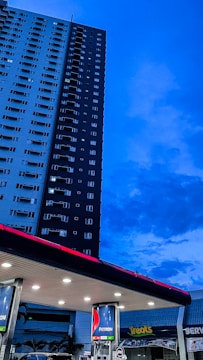 A tall residential building stands adjacent to a gas station, against a backdrop of a deep blue evening sky. The gas station has several fuel pumps visible under a well-lit overhang. Brand signage is prominently displayed on the pumps and the station roof, with a nearby shop with 'Treats' written on its signage.