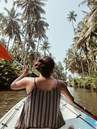 A person is kayaking through a narrow waterway surrounded by tall palm trees on both sides. The individual is holding a paddle with an orange tip and is seated at the back of a white kayak. The atmosphere is serene and tropical, with lush greenery.