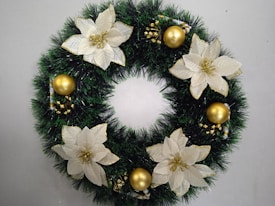 A festive wreath adorned with artificial white poinsettia flowers and golden ornaments, set against a backdrop of lush green foliage.