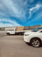 A white pick-up truck carrying a large cardboard box drives on a highway next to another white vehicle. The sky is clear with some wispy clouds, and there is a long, industrial-looking wall in the background.