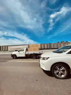 A white pick-up truck carrying a large cardboard box drives on a highway next to another white vehicle. The sky is clear with some wispy clouds, and there is a long, industrial-looking wall in the background.