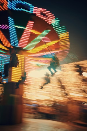 A vibrant amusement park ride with colorful, blurred lights in motion, creating a sense of dynamic movement and excitement. The image captures the swings of a carousel or spinning ride with riders in mid-air, set against a dark evening backdrop.