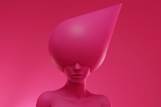 A surreal and stylized image of a person with an oversized, abstract pink headpiece covering their eyes. The entire composition is dominated by shades of pink, creating a striking, modern aesthetic.