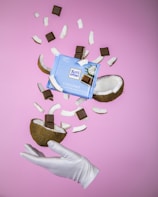 A gloved hand holds a halved coconut with chocolate squares inside. Surrounding the coconut are more chocolate squares and coconut pieces mid-air, against a pink background. A package of Ritter Sport Coconut chocolate is prominently displayed in the middle.