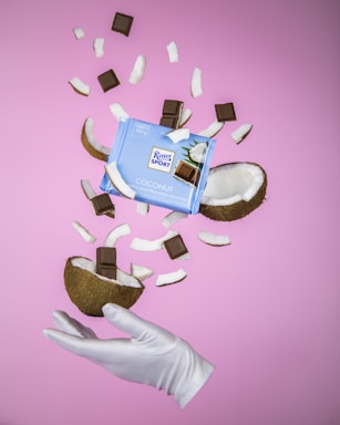 A gloved hand holds a halved coconut with chocolate squares inside. Surrounding the coconut are more chocolate squares and coconut pieces mid-air, against a pink background. A package of Ritter Sport Coconut chocolate is prominently displayed in the middle.