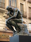 A bronze statue of a muscular man deep in thought, seated on a rock with his chin resting on one hand. Surrounding the statue is a historical building with ornate balconies and detailed stonework.
