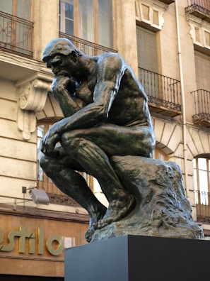 A bronze statue of a muscular man deep in thought, seated on a rock with his chin resting on one hand. Surrounding the statue is a historical building with ornate balconies and detailed stonework.