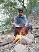 A man in a blue outfit is cooking outdoors over an open fire set among rocks and twigs. Surrounded by lush green trees, he appears focused on managing the pot on the fire. The environment suggests a natural, outdoor setting, possibly in a forest or campsite.