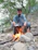 A man in a blue outfit is cooking outdoors over an open fire set among rocks and twigs. Surrounded by lush green trees, he appears focused on managing the pot on the fire. The environment suggests a natural, outdoor setting, possibly in a forest or campsite.
