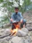 A man in a blue outfit is cooking outdoors over an open fire set among rocks and twigs. Surrounded by lush green trees, he appears focused on managing the pot on the fire. The environment suggests a natural, outdoor setting, possibly in a forest or campsite.