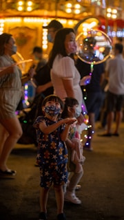 Several people, including a child in star-patterned clothing and a face mask, are gathered at a brightly lit amusement park or fairground. The atmosphere is lively, with lights and colorful decorations. A woman holding a light-up balloon adds to the cheerful environment.