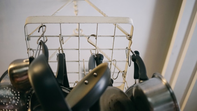 A variety of metal cooking pots and pans hang from hooks attached to a metal wire rack. The rack is suspended from the ceiling, and the image is taken from below, focusing on the kitchen utensils' handles and bases.