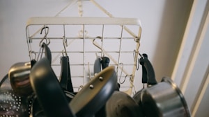 A variety of metal cooking pots and pans hang from hooks attached to a metal wire rack. The rack is suspended from the ceiling, and the image is taken from below, focusing on the kitchen utensils' handles and bases.