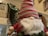 A plush Santa Claus figurine with a large white beard, wearing a red and green patterned hat pulled over its eyes. It is situated on a wooden banister decorated with green garland. The background features a staircase and softly lit Christmas lights.