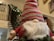 A plush Santa Claus figurine with a large white beard, wearing a red and green patterned hat pulled over its eyes. It is situated on a wooden banister decorated with green garland. The background features a staircase and softly lit Christmas lights.