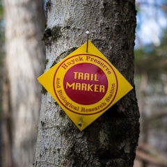 A yellow diamond-shaped trail marker is nailed to the trunk of a tree. The marker has text indicating 'Huyck Preserve Trail Marker and Biological Research Station' in red letters. The background shows a blurred forest scene, suggesting a natural setting.