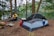 A campsite set in a forested area near a lake, featuring a pitched tent and a person sitting by a campfire. The surroundings are lush with tall trees and a carpet of fallen leaves. The lake in the background provides a serene and peaceful atmosphere.