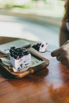 A square slice of blueberry cheesecake is placed on a small wooden tray lined with parchment paper. A hand is holding a fork with a portion of the cake. The cheesecake has a layer of blueberries on top with a creamy middle and a base layer that looks like a crumbly crust. Natural light illuminates the scene, giving it a fresh and inviting appearance.