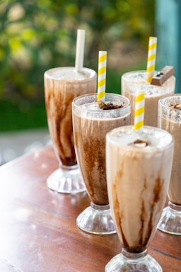 Several tall glasses filled with creamy milkshake topped with chocolate sprinkles and served with striped straws rest on a wooden table. The backdrop includes a soft focus of greenery.