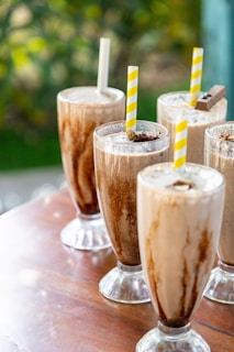 Several tall glasses filled with creamy milkshake topped with chocolate sprinkles and served with striped straws rest on a wooden table. The backdrop includes a soft focus of greenery.