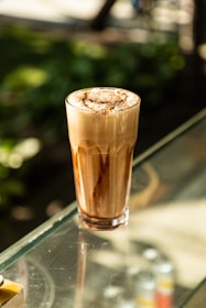 A tall glass of a frothy coffee drink sits on a glass table. The drink has a layered appearance with swirls of chocolate and a creamy froth on top. The background features blurred greenery, suggesting an outdoor setting with natural light.