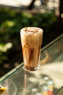 A tall glass of a frothy coffee drink sits on a glass table. The drink has a layered appearance with swirls of chocolate and a creamy froth on top. The background features blurred greenery, suggesting an outdoor setting with natural light.